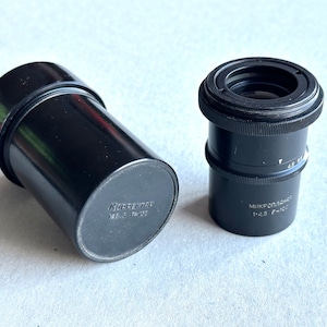 Puede incluir: Dos objetivos de cámara negros, uno con una tapa de lente y el otro con el texto "Mikroplanar 1:4.5 F=100" en el lateral.