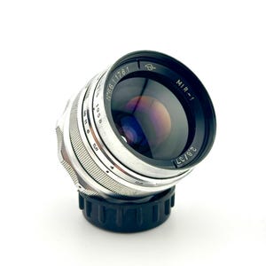 May include: A vintage MIR-1 2.8/37 camera lens. The lens has a black and silver metal body with the text "MIR-1" and "2.8/37" printed on the lens. The lens is angled slightly, showcasing its reflective glass.