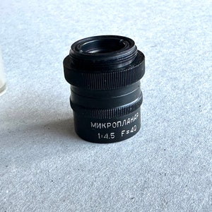 Puede incluir: Un objetivo de cámara negro con el texto "Mikroplanar 1:4.5 F=40" impreso en él.