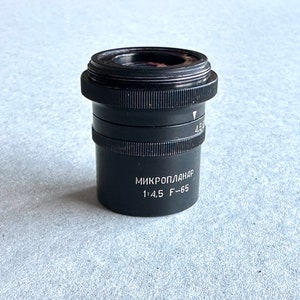 Puede incluir: Un objetivo de cámara negro Mikroplanar 1:4.5 F-65.