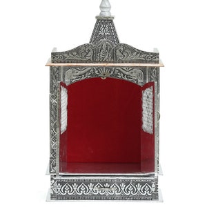 May include: A silver and copper-toned metal temple with intricate floral and geometric designs. The temple has a red velvet interior and a small, pointed roof.
