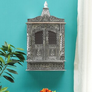 May include: A silver-colored, intricately carved wooden wall-mounted temple with a decorative top and two doors. The temple has a small drawer at the bottom.