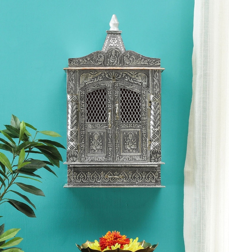May include: A silver-colored, intricately carved wooden wall-mounted temple with a decorative top and two doors. The temple has a small drawer at the bottom.