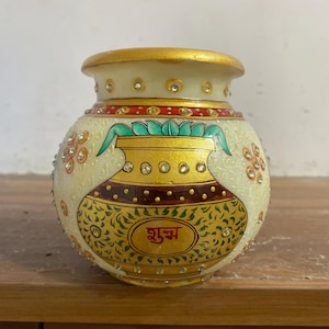 May include: A decorative, off-white pot with gold accents and intricate painted designs. The pot features a gold rim, a central gold and maroon design, and is adorned with small, sparkling embellishments. The word "शुभ" is written in the center of the pot.