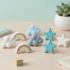 May include: A selection of ceramic drawer pulls shaped like clouds, rainbows, and stars. The pulls are in shades of white, blue, and grey, with rainbow detailing. A wooden block with the letter "A" is also visible.