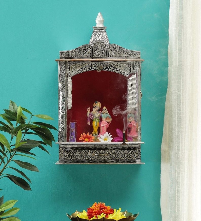 May include: A silver-colored, ornate, wall-mounted temple with a red interior and a glass front. The temple features a small statue of a couple inside. Incense smoke is rising from a stick inside the temple.