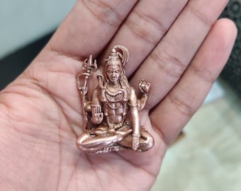May include: A small, copper-colored statue of the Hindu deity Shiva, sitting in a meditative pose with his hands in a mudra. He is holding a trident in his right hand and a small object in his left hand. He is wearing a crown and a necklace.