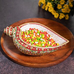 May include: A white marble bowl shaped like a seashell with intricate gold and red painted designs. The bowl is filled with colorful candy, including green, red, yellow, and white candies.