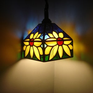 Könnte beinhalten: Eine Hängelampe aus Buntglas mit zwei gelben Blumen mit roten Zentren. Die Blumen sind von blauem und grünem Glas umgeben.