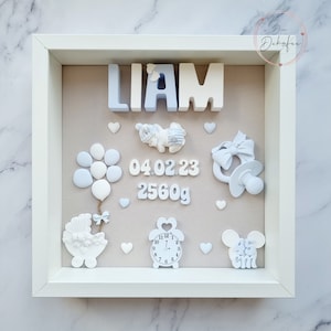 Può includere: Una cornice bianca con sfondo beige e decorazioni blu e bianche. Il nome "LIAM" è scritto in lettere blu e bianche. La data "04.02.23" e il peso "2560g" sono scritti in blu. Un orologio, un ciuccio, un passeggino, un palloncino, un animaletto da bambino e un paio di orecchie di Topolino sono anche inclusi nella cornice.