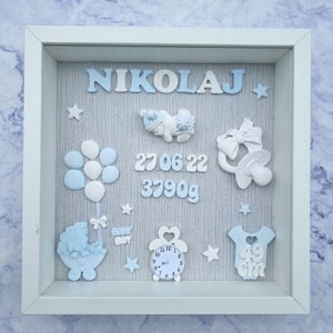 Può includere: Una cornice bianca con sfondo grigio e decorazioni blu e bianche. La cornice include il nome "NIKOLAJ", un bambino che dorme, un ciuccio, palloncini, un passeggino, un orologio e un body con il testo "49 cm". Sono inclusi anche la data "27.06.22" e il peso "3790g".