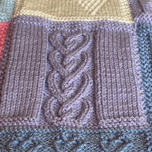 May include: A close-up of a knitted blanket featuring a variety of textures and colours, including purple, blue, and white. The blanket is made up of different squares with different patterns, including a cable knit pattern.