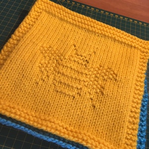 May include: A yellow knitted square with a geometric pattern in the center. The square is bordered with a blue knitted edge.
