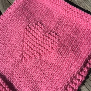 May include: A close-up of a knitted pink blanket featuring a heart design. The blanket is made of soft yarn and has a textured appearance. The heart is created by a different knitting pattern, adding a unique detail.