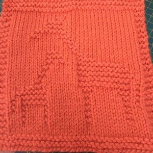 May include: A square, hand-knitted item in a vibrant coral color. The piece features a textured design with geometric shapes, including a stylized star and a horizontal bar. The edges are neatly finished with a ribbed border.