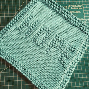 Puede incluir: Una pieza textil tejida de color verde azulado claro, de forma cuadrada, que muestra la palabra HOPE en un patrón de punto texturizado. La pieza tiene un borde en punto bobo y está colocada sobre una base de corte verde con cuadrícula.