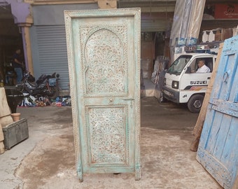 Puede incluir: Una puerta de madera azul claro desgastada con intrincados diseños florales tallados. La puerta tiene un diseño rectangular con un panel central arqueado y un panel rectangular inferior. La puerta está sobre una superficie de hormigón.