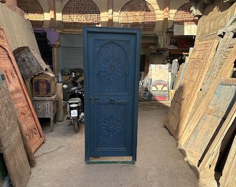 Puede incluir: Una puerta de madera azul oscuro con tallas intrincadas y una manija de metal. La puerta está ubicada en un mercado abarrotado.