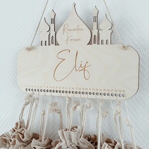 May include: A wooden Ramadan advent calendar with the words "Ramadan Kareem" and "Eliz" engraved on it. The calendar has 30 small fabric bags hanging from it, numbered 1 through 30.