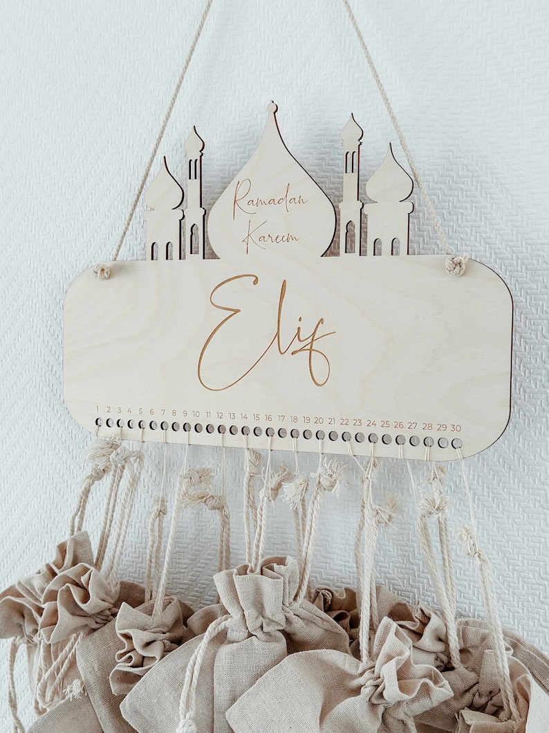 May include: A wooden Ramadan advent calendar with the words "Ramadan Kareem" and "Eliz" engraved on it. The calendar has 30 small fabric bags hanging from it, numbered 1 through 30.