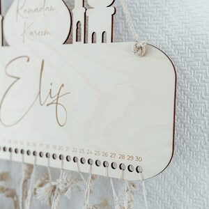 May include: A wooden Ramadan countdown calendar with a mosque design. The calendar has a white background with the words "Ramadan Kareem" and "Elif" written on it. The calendar has 30 numbered pegs for hanging strings.