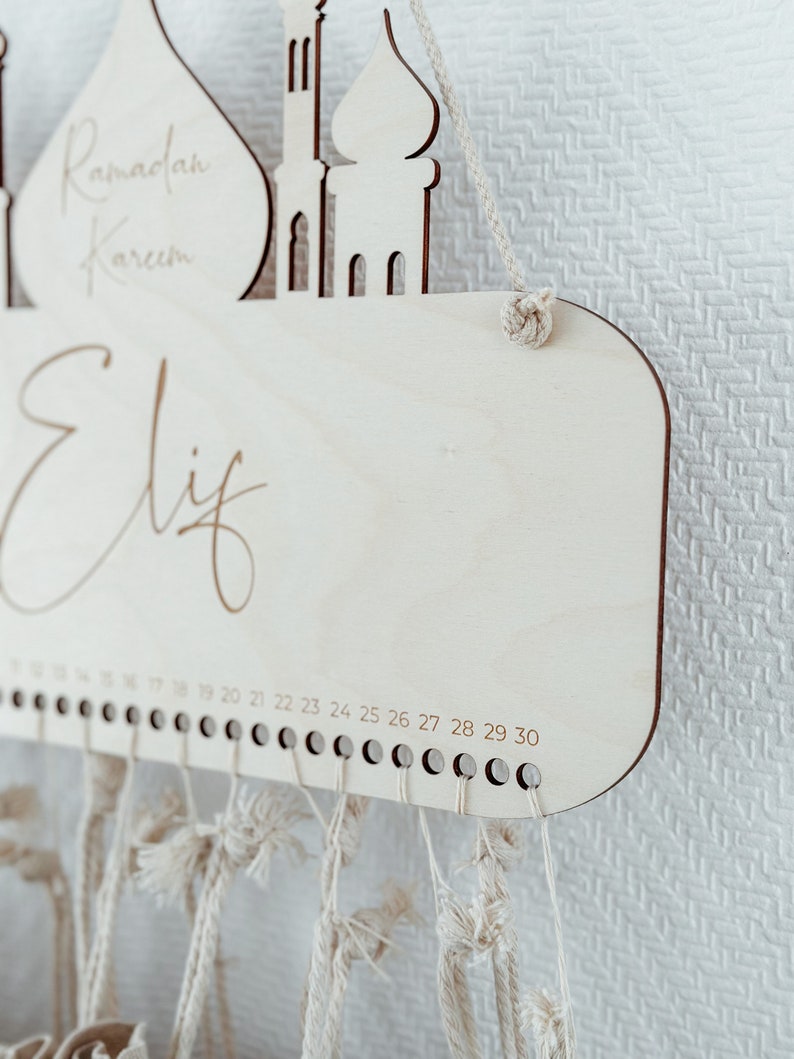 May include: A wooden Ramadan countdown calendar with a mosque design. The calendar has a white background with the words "Ramadan Kareem" and "Elif" written on it. The calendar has 30 numbered pegs for hanging strings.