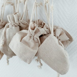 May include: A set of small, beige, fabric drawstring bags hanging from a wooden rack. The bags are made of a natural, linen-like material and have a rustic, handmade look.