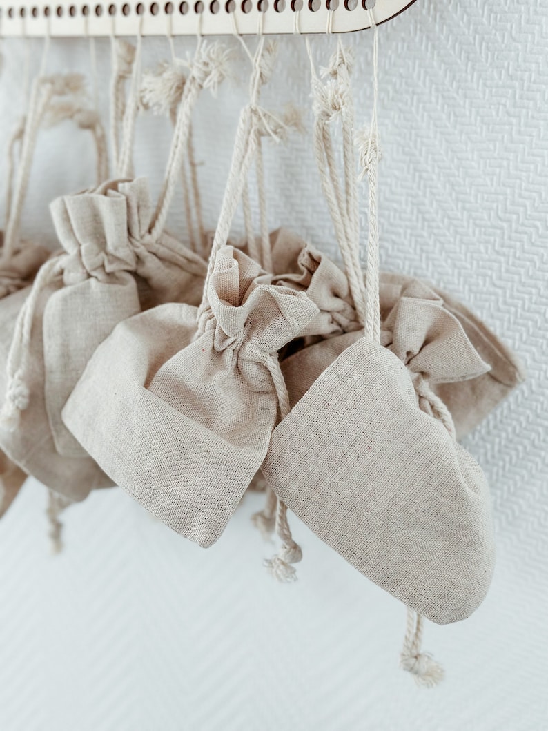 May include: A set of small, beige, fabric drawstring bags hanging from a wooden rack. The bags are made of a natural, linen-like material and have a rustic, handmade look.