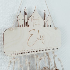 May include: Wooden Ramadan advent calendar with a mosque design and the text "Ramadan Kareem" and "Elif". The calendar has 30 small fabric bags hanging from it.