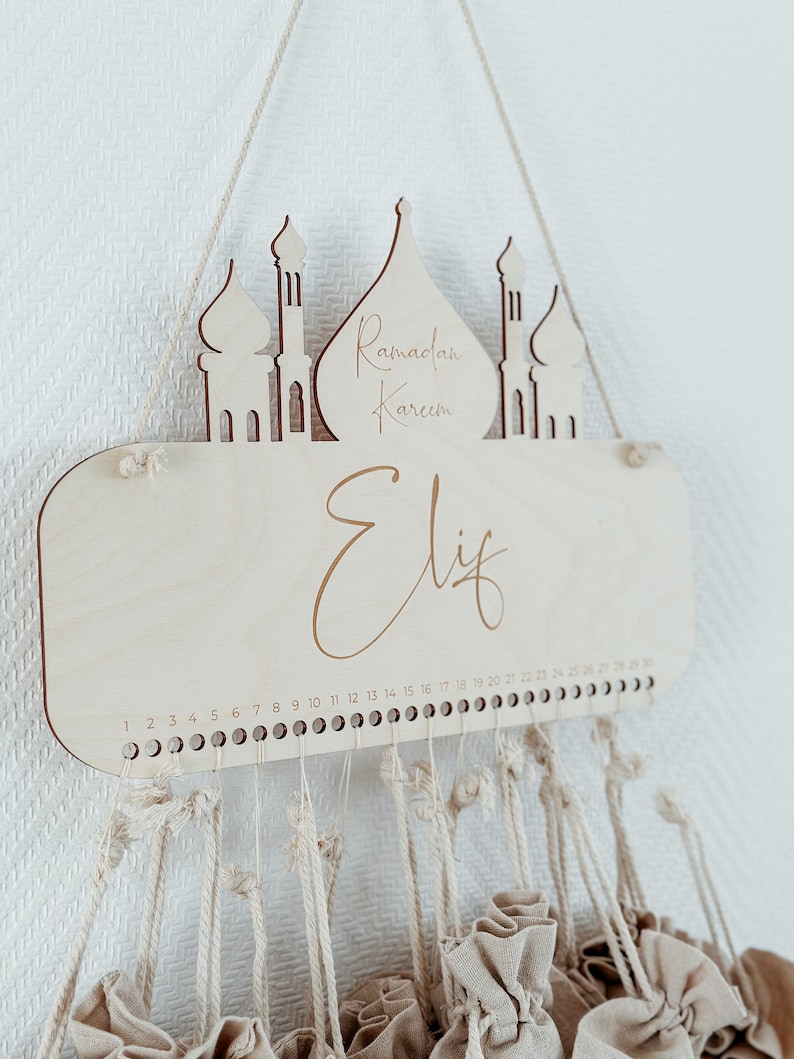 May include: Wooden Ramadan advent calendar with a mosque design and the text "Ramadan Kareem" and "Elif". The calendar has 30 small fabric bags hanging from it.