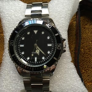 May include: A silver-toned wristwatch with a black dial and a rotating bezel. The watch features luminous markers and hands, and a stainless steel band. The watch is displayed on a white cushion.