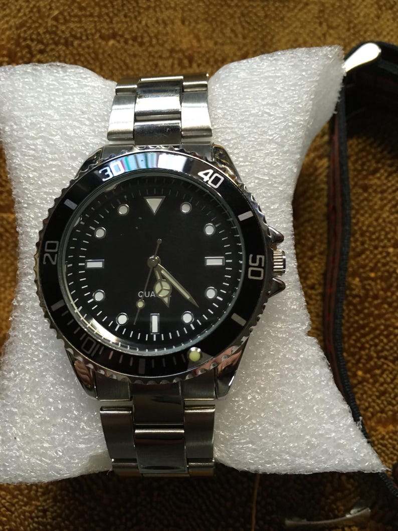 May include: A silver-toned wristwatch with a black dial and a rotating bezel. The watch features luminous markers and hands, and a stainless steel band. The watch is displayed on a white cushion.