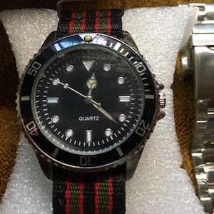 May include: A black quartz watch with a black bezel and a black dial. The watch has a black, red, and green striped band. A silver watch band is next to the watch.