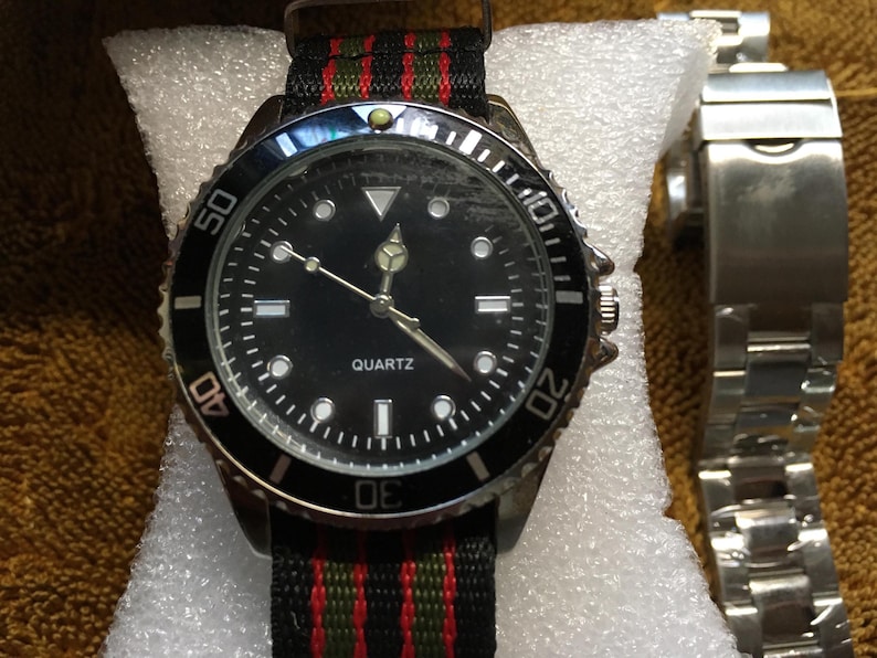 May include: A black quartz watch with a black bezel and a black dial. The watch has a black, red, and green striped band. A silver watch band is next to the watch.