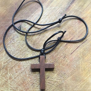 May include: A wooden cross pendant necklace with a black cord. The cross is a medium brown color and has a simple, classic design. The cord is adjustable, allowing for a comfortable fit. The necklace is displayed on a wooden surface.