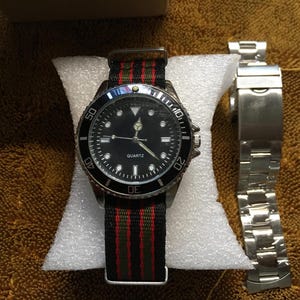 May include: A black quartz watch with a black bezel and a black dial, featuring white hour markers and hands. The watch has a striped green, red, and black nylon band. A separate silver metal watch band is also present.