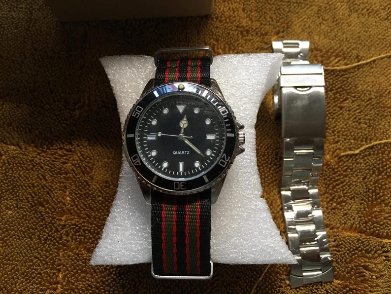 May include: A black quartz watch with a black bezel and a black dial, featuring white hour markers and hands. The watch has a striped green, red, and black nylon band. A separate silver metal watch band is also present.