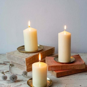 May include: Three white pillar candles with flames burning on gold metal candle holders. The candles are on top of a stack of books.