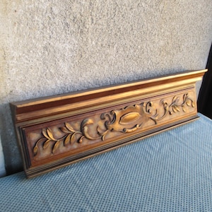Puede incluir: Una moldura decorativa rectangular de madera con intrincados tallados florales y de hojas doradas. La moldura tiene una base marrón oscuro y un borde superior dorado. La pieza es probablemente para diseño de interiores o detalles arquitectónicos.