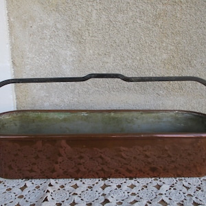 May include: A rectangular copper planter with a dark metal handle. The planter has a brown exterior and a green interior. The handle arches over the top of the planter. The planter is sitting on a white lace doily.
