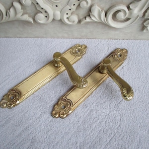 Puede incluir: Dos manillas de puerta doradas adornadas con intrincados diseños florales. Las manillas son de metal y tienen un acabado pulido.
