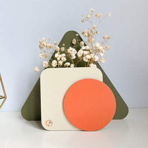 May include: A white geometric vase with a large orange circle on the front, sitting on a green triangular base. The vase is filled with dried white flowers.
