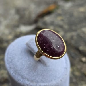 Może przedstawiać: Złoty pierścionek z dużym, owalnym, ciemnofioletowym kamieniem szlachetnym. Kamień szlachetny jest osadzony w złotej oprawie i ma nakrapiany wygląd. Pierścionek jest prezentowany na jasnoszarej powierzchni, prezentując wzór biżuterii.