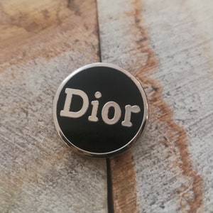 Peut inclure: Une épingle circulaire noire et argentée avec le mot "Dior" en lettres blanches.