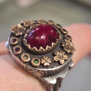 May include: Close-up of a handcrafted ring featuring a large, oval, deep red gemstone at the center. The ring has a silver band with a textured design and is adorned with small gold-colored flowers and gemstones.