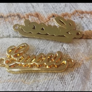 Może przedstawiać: Dwa złote metalowe breloki z napisem "Gucci" napisanym kursywą. Breloki są wycięte i mają otwór u góry do przymocowania do naszyjnika lub bransoletki.