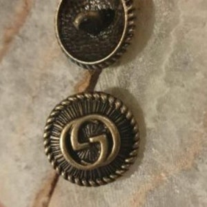May include: Two gold-toned metal buttons with a raised interlocking G logo design. The buttons have a textured, circular design around the edge.