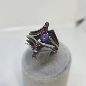 May include: A silver-toned ring with an intricate, layered design. The ring features a large, pear-shaped purple gemstone and several smaller pink and clear gemstones. The ring is displayed on a gray surface.