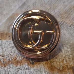 Op de afbeelding: Een goudkleurige, ronde knoop met een verhoogd dubbel-G-logo in het midden. De knoop heeft een glanzende, metallic afwerking en een geribbelde rand. De knoop ligt op een getextureerd, lichtgekleurd oppervlak.