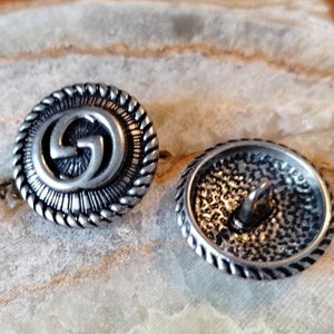 Pode incluir: Dois botões prateados com um design de corda ao redor da borda. Um botão apresenta um design estilizado de 'G' entrelaçadas no centro. O outro botão tem uma superfície texturizada.